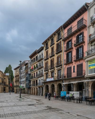 A wide, paved public square lined with multi-story buildings featuring arched ground-floor arcades and balconies, under an overcast sky.