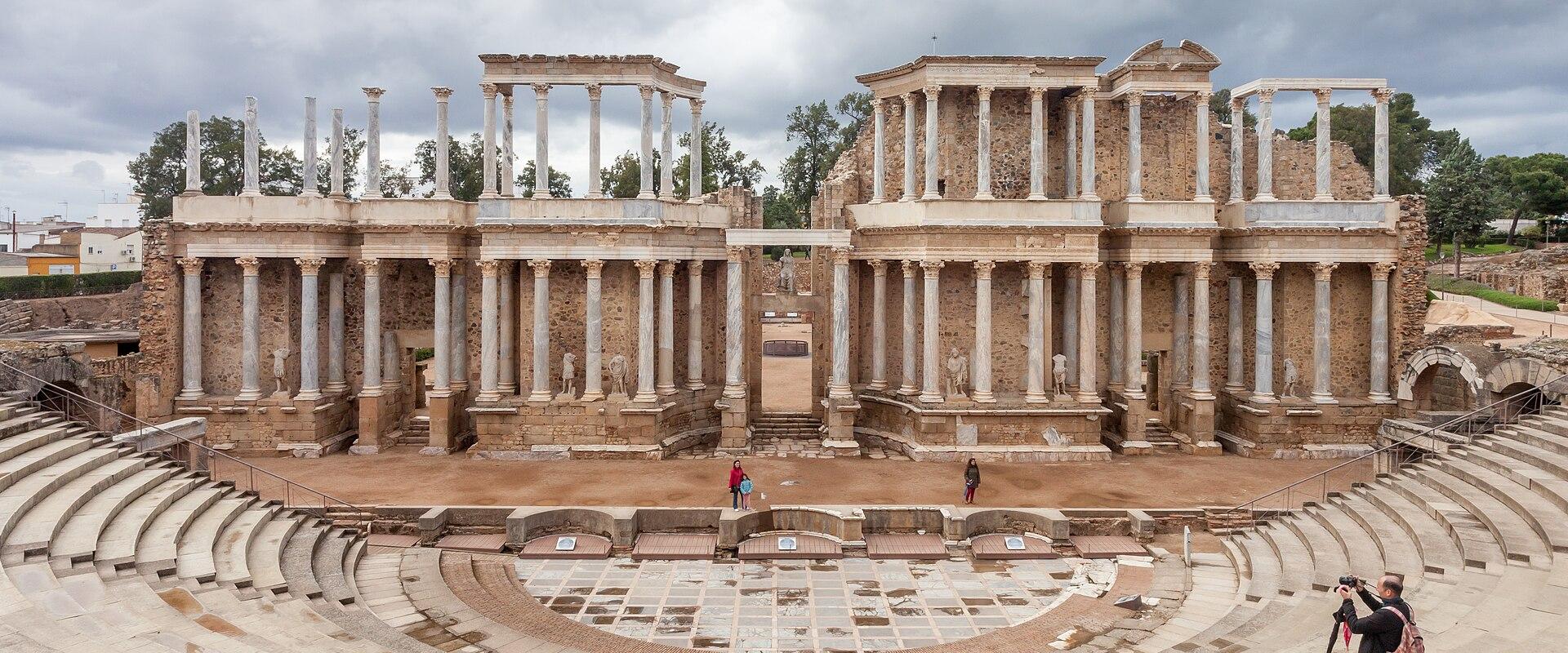 A wide-angle view of the Roman Theatre of Mérida showing the semi-circular seating area (cavea), the orchestra, and the reconstructed two-story stage building (scaenae frons) with its columns and arches under a cloudy sky.