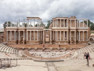 A wide-angle view of the Roman Theatre of Mérida showing the semi-circular seating area (cavea), the orchestra, and the reconstructed two-story stage building (scaenae frons) with its columns and arches under a cloudy sky.
