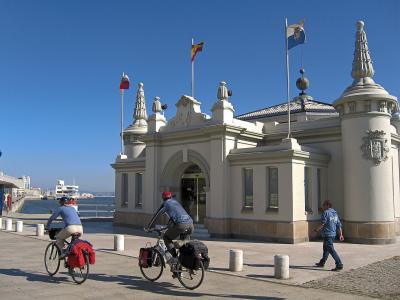 A white, ornate building with turrets and flags (Spanish, Cantabrian, and Santander municipal) stands by a harbor, with cyclists passing on a paved promenade in the foreground.