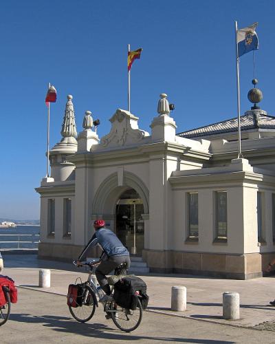 A white, ornate building with turrets and flags (Spanish, Cantabrian, and Santander municipal) stands by a harbor, with cyclists passing on a paved promenade in the foreground.