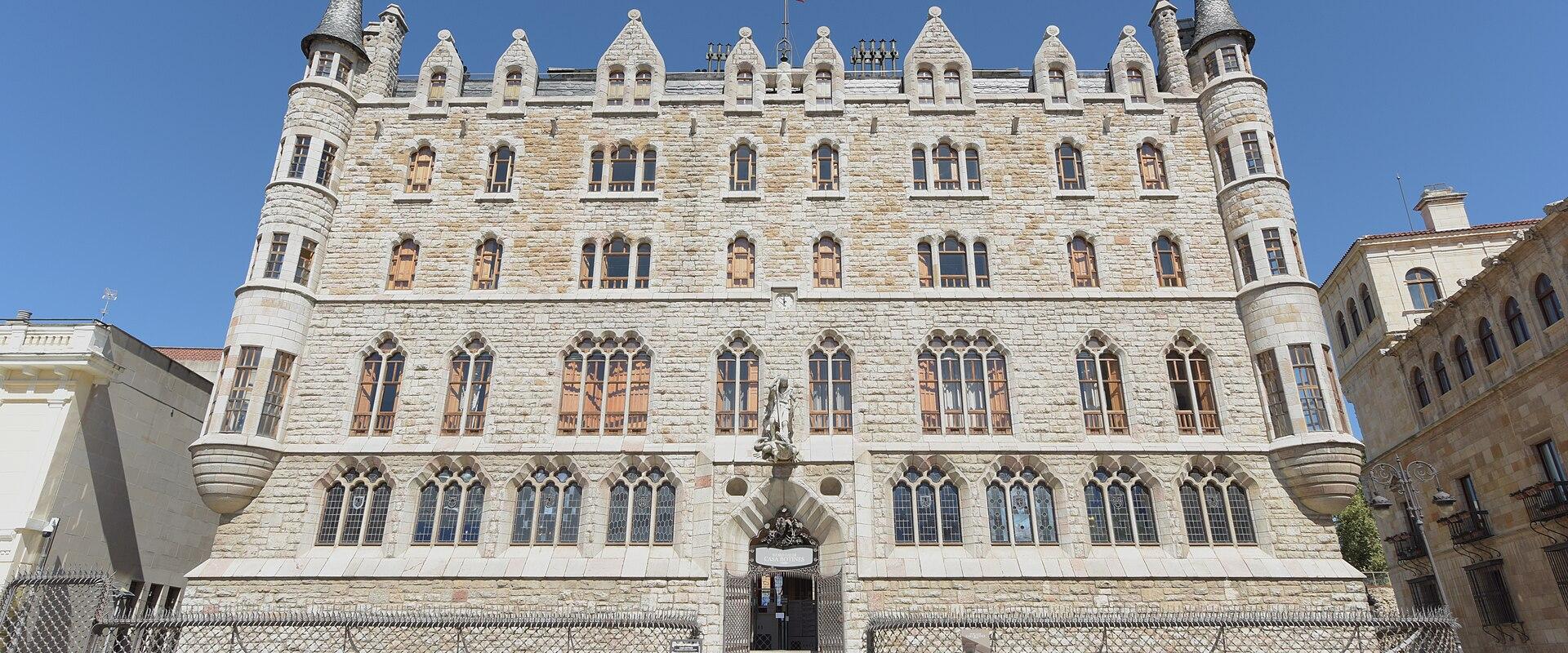 A large, ornate stone building with Gothic Revival and Neo-Gothic features, including corner turrets with conical roofs, arched windows, and a central entrance under a statue; a Spanish flag flies atop the roof; the building is situated in an open pl