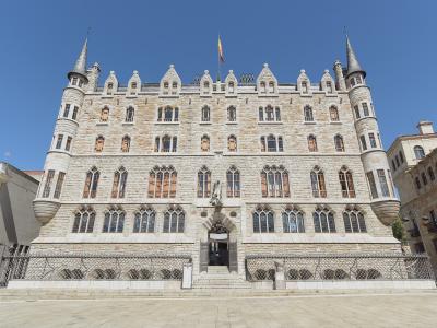 A large, ornate stone building with Gothic Revival and Neo-Gothic features, including corner turrets with conical roofs, arched windows, and a central entrance under a statue; a Spanish flag flies atop the roof; the building is situated in an open pl