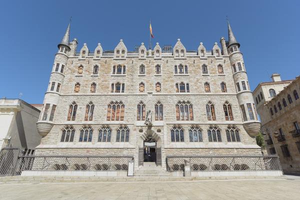 A large, ornate stone building with Gothic Revival and Neo-Gothic features, including corner turrets with conical roofs, arched windows, and a central entrance under a statue; a Spanish flag flies atop the roof; the building is situated in an open pl