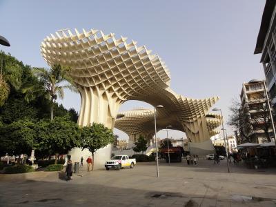 A large, modern wooden lattice structure with mushroom-like canopies supported by curved columns, situated in a paved public plaza with trees, pedestrians, and a white utility vehicle.