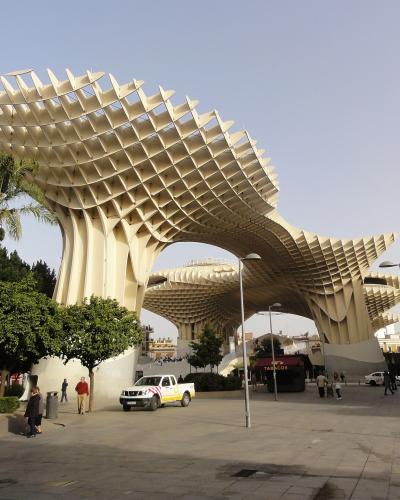 A large, modern wooden lattice structure with mushroom-like canopies supported by curved columns, situated in a paved public plaza with trees, pedestrians, and a white utility vehicle.