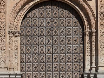 A close-up of an ornate, arched stone doorway with a large, studded wooden door, featuring elaborate Plateresque-style carvings including a coat of arms, cherubs, and busts.