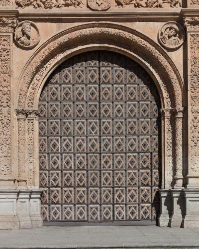 A close-up of an ornate, arched stone doorway with a large, studded wooden door, featuring elaborate Plateresque-style carvings including a coat of arms, cherubs, and busts.