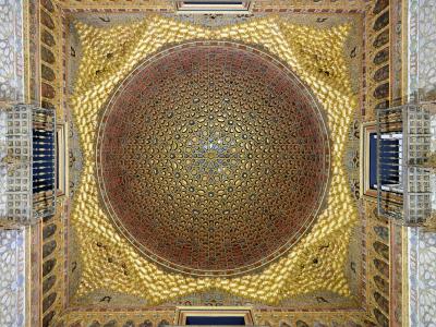 An ornate, gilded wooden ceiling with intricate geometric and floral patterns, featuring a central dome-like structure and four projecting balconies, characteristic of Mudéjar architecture.