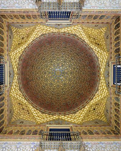 An ornate, gilded wooden ceiling with intricate geometric and floral patterns, featuring a central dome-like structure and four projecting balconies, characteristic of Mudéjar architecture.