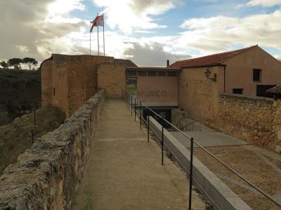 The image shows the exterior of a stone fortress-like structure with a modern entrance ramp and a sign reading "MUSEO" on a beige wall, with flags flying above.