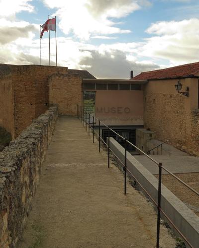 The image shows the exterior of a stone fortress-like structure with a modern entrance ramp and a sign reading "MUSEO" on a beige wall, with flags flying above.