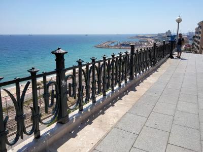 A paved promenade with a decorative black metal railing overlooks a beach, railway tracks, and a large harbor with ships and cranes in the distance under a clear blue sky.