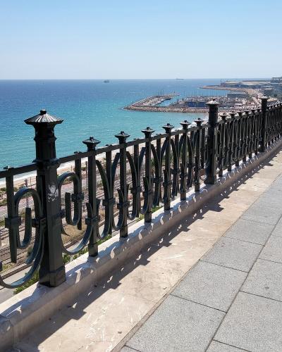 A paved promenade with a decorative black metal railing overlooks a beach, railway tracks, and a large harbor with ships and cranes in the distance under a clear blue sky.