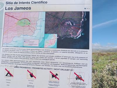 A large informational signpost outdoors displaying a map, aerial photo, and multilingual text identifying the location as "Los Jameos" under the heading "Sitio de Interés Científico," with prohibited activity icons at the bottom.