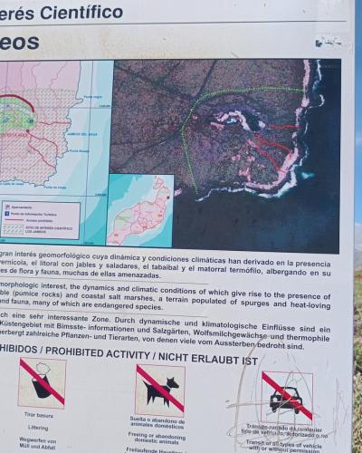 A large informational signpost outdoors displaying a map, aerial photo, and multilingual text identifying the location as "Los Jameos" under the heading "Sitio de Interés Científico," with prohibited activity icons at the bottom.