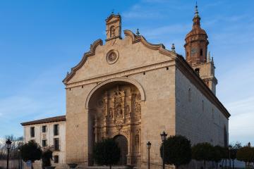A large stone church with an ornate arched entrance and a tall, tiled bell tower stands in a town square under a blue sky.
