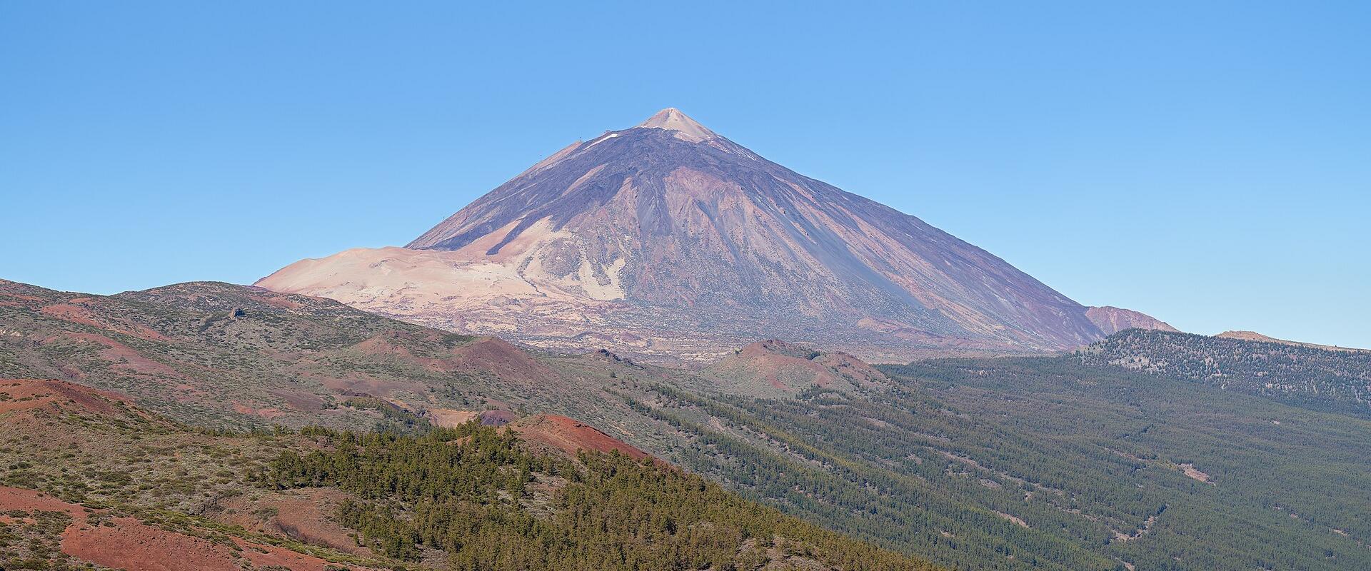 A large, conical stratovolcano with a distinct crater at its peak, viewed from a distance across a foreground of reddish volcanic soil and green pine forests under a clear blue sky.
