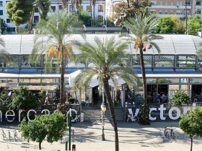 A modern, glass-roofed market building with outdoor seating, palm trees, and large white letters spelling "mercado victoria" on the ground in front.