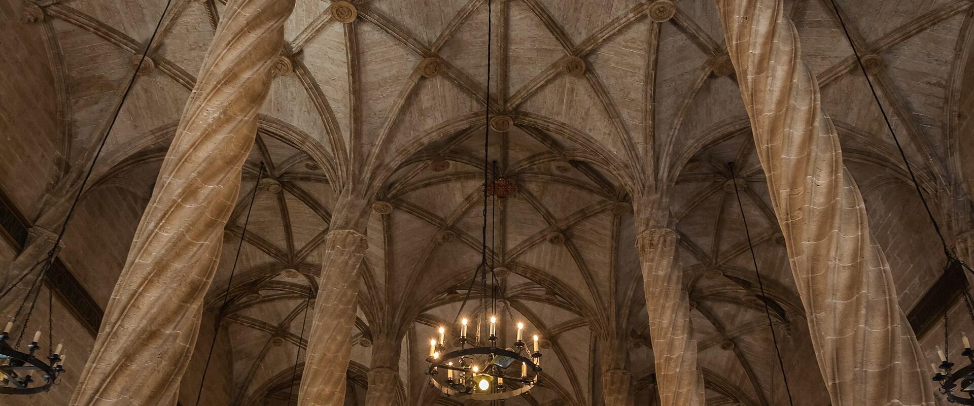 The interior of a Gothic hall featuring tall, twisted stone columns supporting a complex ribbed vault ceiling, with several large chandeliers hanging from above and visitors walking on the floor.