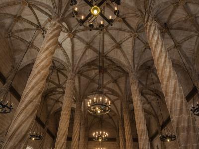 The interior of a Gothic hall featuring tall, twisted stone columns supporting a complex ribbed vault ceiling, with several large chandeliers hanging from above and visitors walking on the floor.