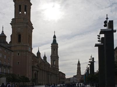 A large baroque cathedral with two prominent bell towers and a green copper spire, situated next to a modern plaza with a reflecting pool and contemporary black light fixtures.