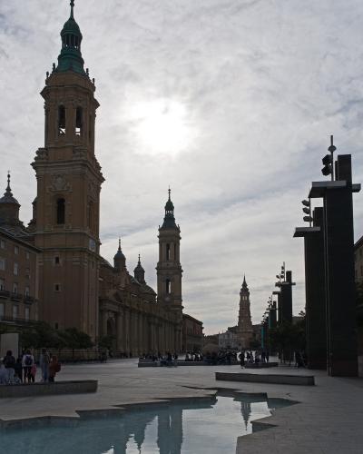 A large baroque cathedral with two prominent bell towers and a green copper spire, situated next to a modern plaza with a reflecting pool and contemporary black light fixtures.